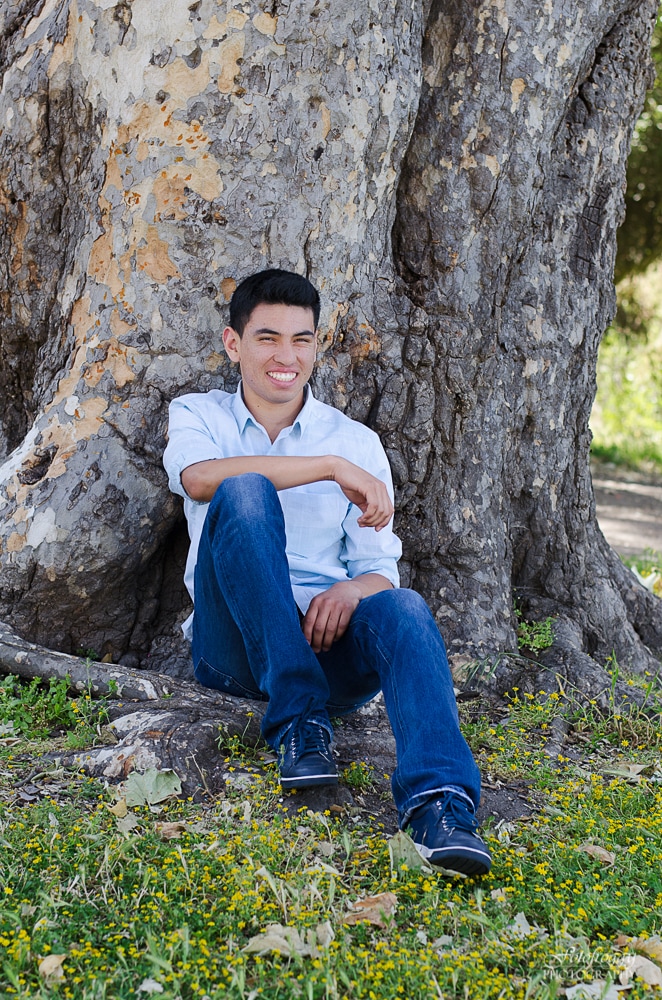 Senior boy sitting at base of large tree laughing.