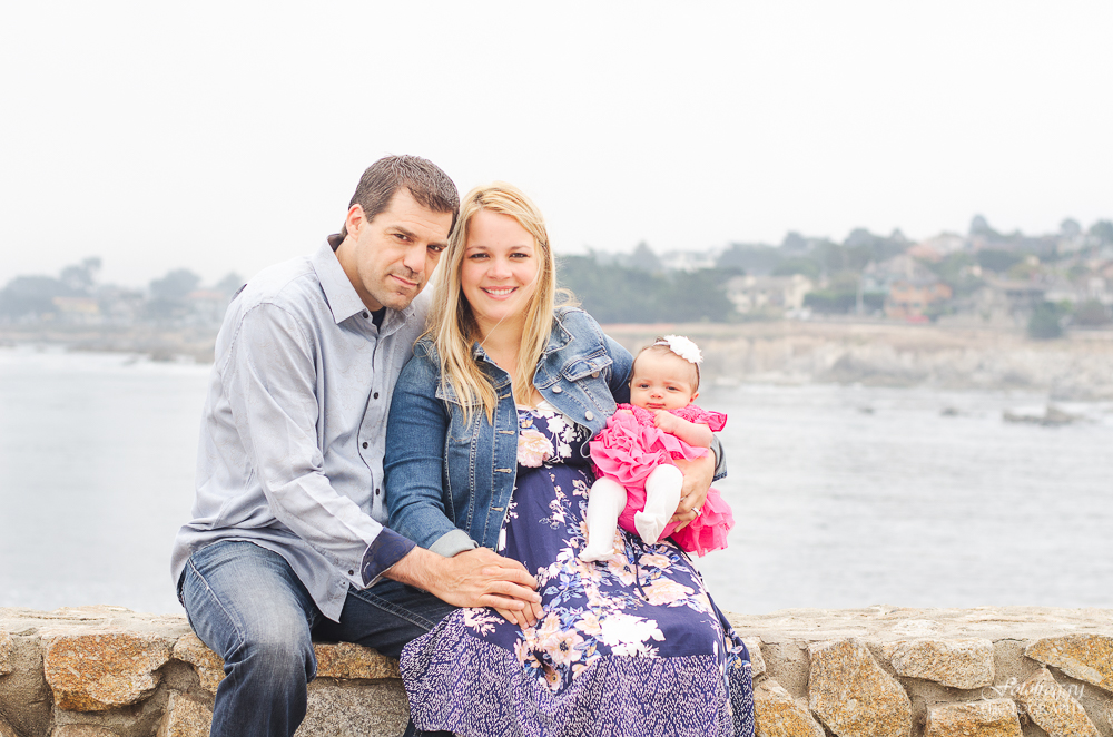Cute couple with adorable babygirl in pink ruffles sitting on rock wall Monterey Bay Ca
